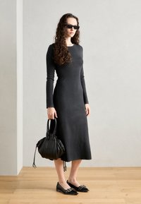 Long-sleeved, fitted, dark gray knit dress, flat black shoes with embellishments, and a black handbag with two handles. Plain background.