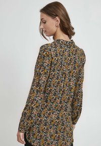 Květinová halenka s dlouhým rukávem z tmavé látky se žlutými, hnědými a modrými vzory. Má zapínání na knoflíky na zadní straně a volný střih s měkkou texturou.