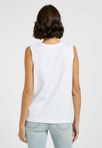 Top sin mangas blanco en tejido suave con escote redondo, detalles de borde sin acabar y un corte holgado, combinado con jeans de un azul claro.