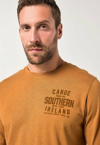 Langsleeve t-shirt in mosterdgeel, gemaakt van zachte stof, met een bedrukte opdruk van "Canoe Trail Co." en "Zuid-Ierland" in donkerbruin.