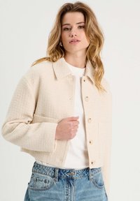 Gekürzte cremefarbene Jacke mit Struktur, Kragen, Vorderknöpfen und zwei Taschen. Über einem weißen Hemd getragen und mit blauen Jeans kombiniert.