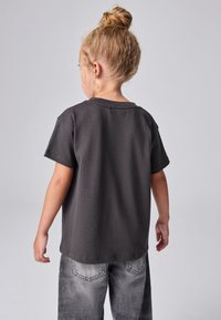 Dieťa so svetlohnedými vlasmi zviazanými do drdola, oblečené v obyčajnom čiernom tričku a vyblednutých sivých džínsach, stojace chrbtom k prednej strane na sivom pozadí.