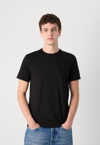 Zwarte katoenen t-shirt met ronde hals en korte mouwen, met een subtiele logo op de borst. Draagt het met lichtblauwe jeans.