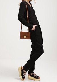 Haut noir boutonné, pantalon noir assorti et sandales à plateforme avec des brides croisées. Sac bandoulière marron avec des éléments en métal doré.
