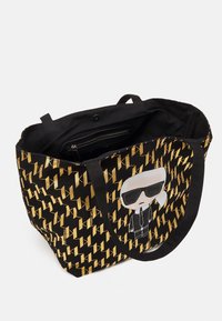 Bolsa de mano negra con un patrón geométrico dorado, una figura de dibujos animados con gafas de sol y asas negras resistentes. Bolsillo interior con cremallera.