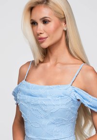 Femme blonde aux longs cheveux portant une robe bleue claire décolletée texturée et de petites boucles d’oreilles rondes, regardant sur le côté.