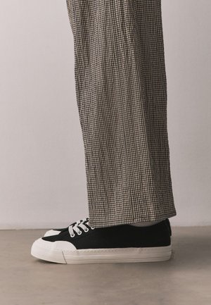 Vue sur toute la jambe d'une personne portant des baskets en toile noires et blanches avec des lacets blancs et un pantalon large marron à carreaux.