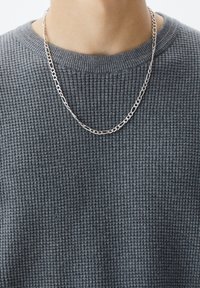 Silberne Kettenhalskette mit ineinandergreifenden Gliedern, getragen über einem strukturierten, dunkelgrauen, gerippten Pullover.