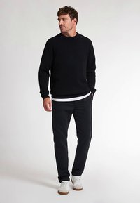 Schwarzer Pullover aus weichem Stoff mit Rundhalsausschnitt und gerippten Bündchen, kombiniert mit schwarzer Hose und weißen Sneakers mit hellbraunen Sohlen.