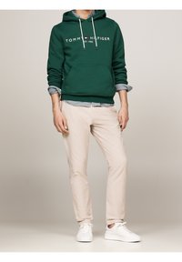 Zelená mikina s kapucí s přední kapsou a bílým logem, navrstvená přes kostkovanou košili. Světle béžové kalhoty a bílé tenisky doplňují outfit.