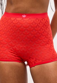 Κοντινό πλάνο ατόμου που φορεί κόκκινα σορτς με ανάγλυφο επαναλαμβανόμενο μοτίβο λογότυπου Adidas και μικρό λογότυπο στη μέση της ζώνης.