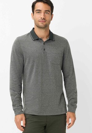 Man wearing grey long-sleeve polo shirt with black collar and dark green pants standing against white background.