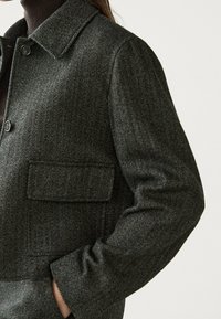Kurtka w ciemnoszarym kolorze w wzór jodełki, z kołnierzem, zapięciem na guziki z przodu i kieszenią na piersi dla dodatkowego akcentu. Miękki, teksturowany materiał.