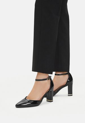 Czarne lakierowane skórzane czółenka na słupku z ostrym noskiem, paskiem wokół kostki i złotym akcentem na pięcie, zestawione z dopasowanymi czarnymi spodniami.