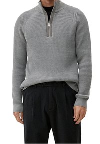 Homme portant un pull en maille côtelée gris clair avec un col à demi-fermeture éclair, par-dessus une chemise noire et un pantalon noir sur mesure.