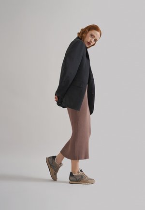 Schwarzes Blazer, gerippter brauner Midirock und grau-braune Sneaker mit Fellfutter und strukturierten Akzenten. Model in Seitenansicht vor einem neutralen Hintergrund.