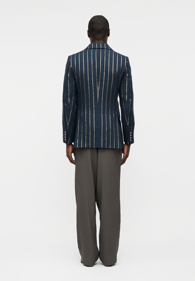 Άνδρας που στέκεται με την πλάτη προς τα έξω, φοράει μπλε σκούρο σακάκι με λευκές κάθετες ρίγες και άνετο γκρι παντελόνι, μπροστά σε απλό φόντο.