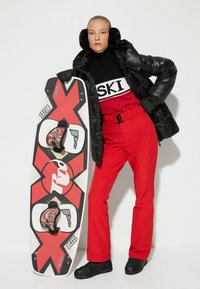 Chaqueta acolchada negra, suéter rojo con el texto "SKI", pantalones rojos, zapatos negros, sosteniendo una tabla de snowboard roja y blanca con diseño gráfico.