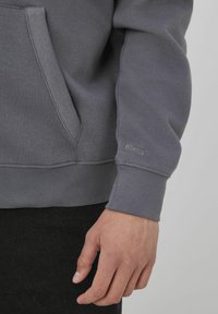 Sweatshirt gris avec une poche kangourou, des poignets côtelés et un logo subtil sur la manche. Texture lisse avec une coupe décontractée.