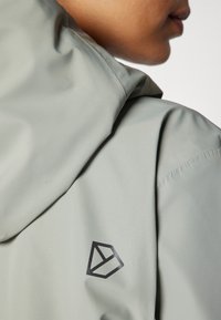 Veste vert clair avec une texture lisse, présentant un logo géométrique noir sur la capuche et un tissu doux avec des coutures minimales.