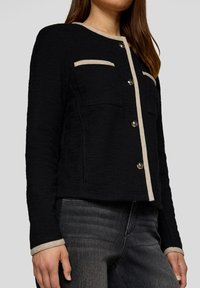 Schwarze strukturierte Jacke mit beigem Besatz, runder Halslinie und Knopfschluss. Verfügt über zwei Brusttaschen und lange Ärmel mit kontrastierenden Bündchen.