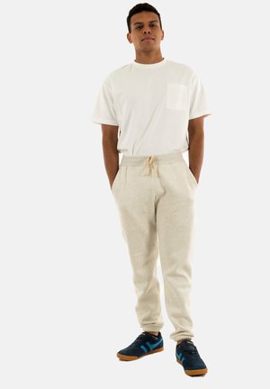 Conjunto de hombre que incluye una camiseta de algodón blanca con un bolsillo en el pecho, pantalones de chándal beige claros y zapatillas navy con detalles azules y suelas marrones.