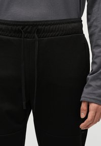 Άνδρας που φοράει μαύρο παντελόνι τζόγκερ με ελαστική μέση και κορδόνια, συνδυασμένο με γκρι μακρυμάνικο πουκάμισο, με το χέρι ακουμπισμένο στο πλάι.