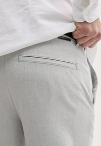 Pantalons texturés gris clair avec une poche arrière et une étiquette près de la taille, partiellement cachée par une chemise blanche.