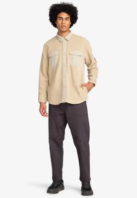 Giacca in pile beige con chiusura a bottoni, due tasche sul petto e colletto a contrasto. Indossata con pantaloni scuri e scarpe nere.