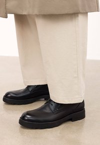 Zapatos de cuero negro con detalles texturizados, suelas de goma negra y cordones negros, combinados con pantalones de corte relajado en color crema.