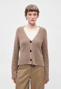 Cardigã castanho tricotado com decote em V, apresentando três botões pretos e mangas longas. Usado sobre uma camisa creme e combinado com calças bejas.