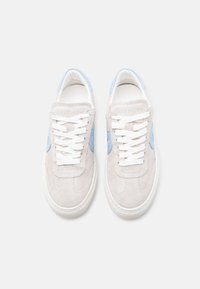 Ζεύγος ανοιχτόχρωμων μπεζ σουέτ αθλητικών παπουτσιών με λευκά κορδόνια και γαλάζιες λεπτομέρειες, απεικονιζόμενα από ψηλά σε λευκό φόντο.