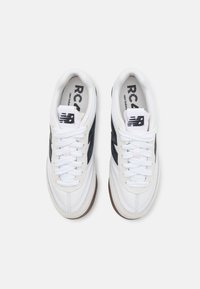 Witte sportieve schoenen met zwarte accenten, voorzien van een textiel leren bovenwerk, ronde neus en platte veters, geplaatst tegen een lichte achtergrond.