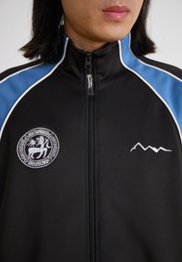 Black zip-up jacket with blue shoulder stripes, featuring a round embroidered logo and a mountain design accent in white. Stretch material.