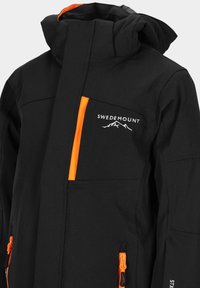 Veste noire avec un accent orange sur la poche avant. Présente le logo "SWEDEMOUNT" et plusieurs poches zippées. Texture lisse.