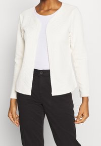 Weißer strukturierter Cardigan mit Zickzackmuster, offenem Vorderteil und langen Ärmeln. Kombiniert mit einem hellen Oberteil und einer dunklen Hose.