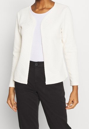 Witte getextureerde cardigan met een zigzagpatroon, open voorkant en lange mouwen. Gecombineerd met een licht shirt en donkere broek.