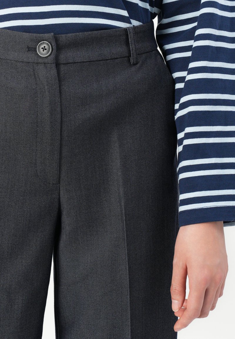 Pantaloni grigi su misura realizzati in tessuto texturizzato con chiusura a bottone, davanti piatto e tasche laterali. Maglietta a righe blu e bianche visibile.