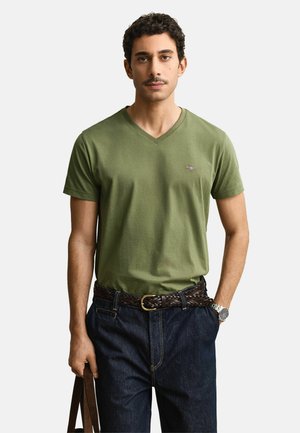 Homme portant un T-shirt col en V vert olive, un jean foncé avec une ceinture tressée marron, une montre-bracelet argentée et tenant la poignée d’un sac en cuir marron.