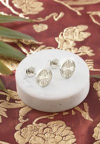 Silverfärgade studs med cirkulär design som har geometriska mönster, visade på en vit marmorbas mot en texturerad röd och guld bakgrund.