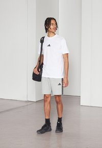 Λευκό κοντομάνικο μπλουζάκι με μαύρο λογότυπο adidas, συνδυασμένο με γκρίζα σορτς με μαύρες ρίγες στο πλάι. Μαύρα αθλητικά παπούτσια και σκούρα τσάντα.