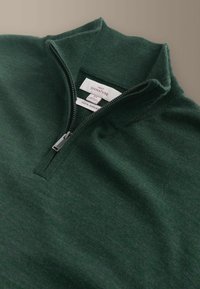 Mörkgrön polotröja i merinoull med kvartszip, utrustad med ståkrage och mjuk, slät textur. Etiketten visar 100 % merinoull.