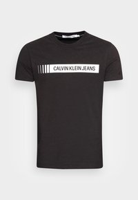 Černé tričko s krátkým rukávem a bílým obdélníkovým logem a textem "Calvin Klein Jeans" přes hrudník.