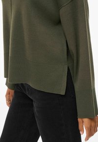 Pull tricot olive en maille avec des poignets et un ourlet côtelés, des fentes sur les côtés et une coupe décontractée, associé à un pantalon noir fuselé.