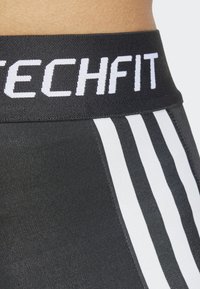 Crne atletske tajice s širokim crnim pojasom na kojem je bijeli tekst "TECHFIT". Dvije vertikalne bijele pruge duž strana. Glatka tekstura.