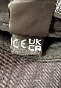 Étiquette de vêtement montrant les marquages de conformité CE et UKCA cousus sur un tissu foncé avec des coutures visibles et un patch en cuir plié au-dessus.