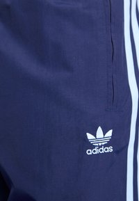 Tengerészkék sportnadrág világoskék oldalsávokkal és hímzett fehér Adidas logóval a bal oldalon. Könnyű anyag, sima textúra.