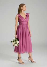Robe en chiffon rose avec un décolleté en V, accent floral sur l'épaule, jupe plissée et sandales à talons hauts argentées, tenant un bouquet de fleurs.