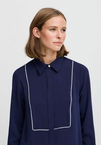 Camicia blu navy con colletto, decorata con ricami bianchi a scallop sulla parte anteriore. Tessuto morbido con chiusura a bottone al collo.