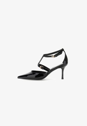 Scarpa con tacco alto in pelle patent nera, punta affusolata, cinturini alla caviglia, dettagli in metallo dorato e tacco a stiletto.